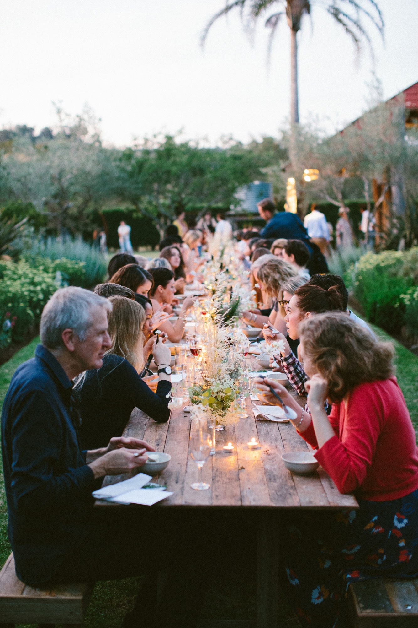 Guests sharing a family-style dinner at a long wooden table in a garden at dusk